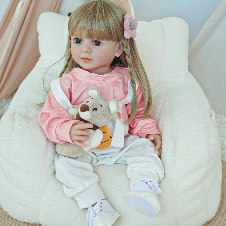 Reborn toddler doll sitting in a soft chair, dressed in pink and white overalls, holding a small plush toy.