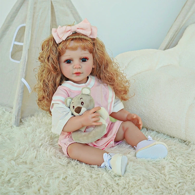 Reborn toddler doll sitting on a soft white surface, holding a plush bear, wearing a pink dress and bow.