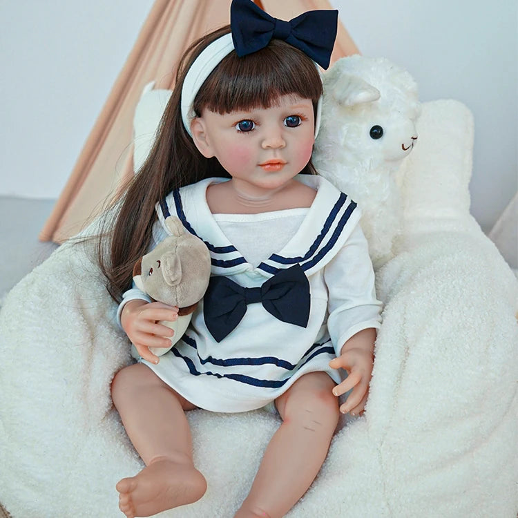 Reborn toddler doll sitting on a soft chair, wearing a sailor dress and holding a teddy bear, with a cute navy blue bow in her hair.