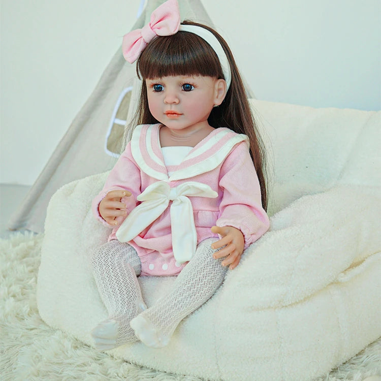 Reborn toddler doll sitting on a plush chair, wearing a pink dress with a white bow and gray tights, holding a teddy bear.