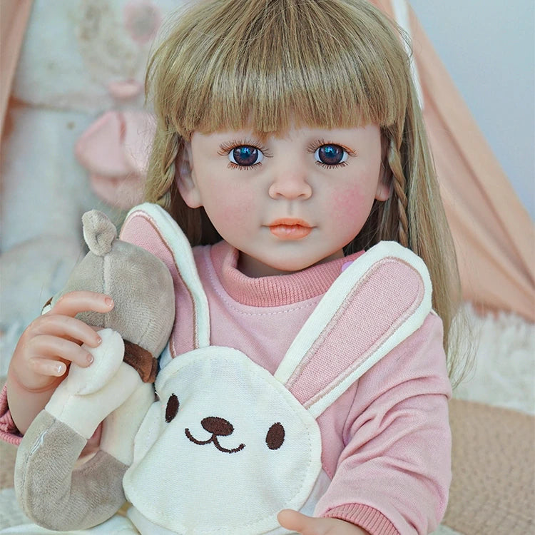 Close-up of reborn toddler doll holding a teddy bear, wearing white overalls with a bunny design and a pink sweater.