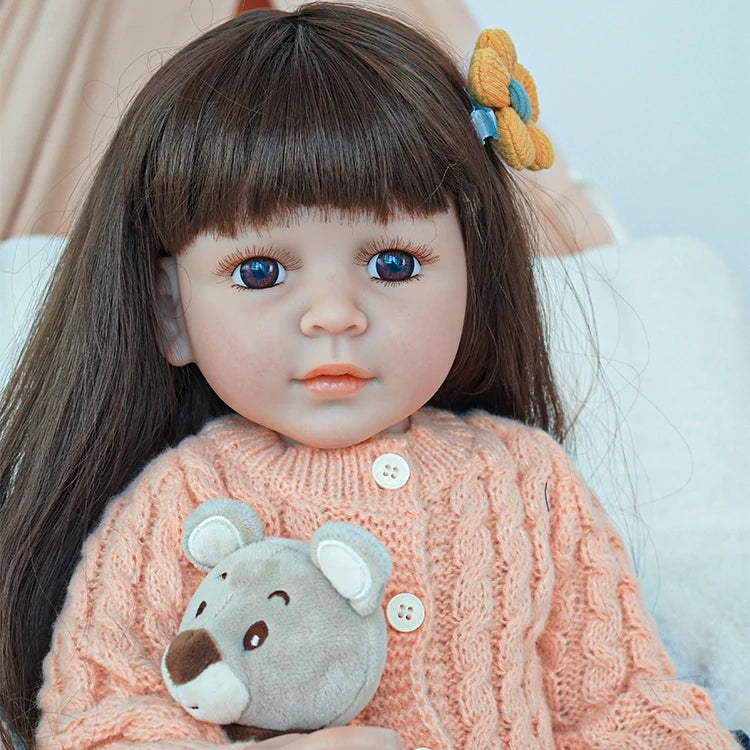 Close-up of reborn toddler doll with long brown hair, dressed in a peach knit sweater and holding a plush bear.