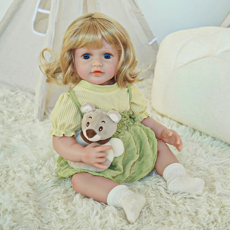 Reborn toddler doll sitting on a soft rug, dressed in a green dress, holding a small teddy bear, with white socks.