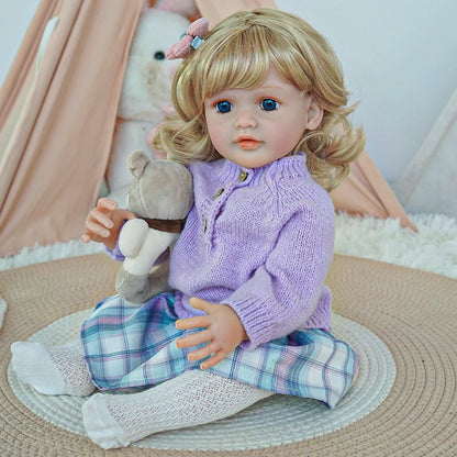 Reborn toddler doll sitting on a soft rug, dressed in a purple sweater and plaid pants, holding a teddy bear.