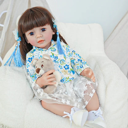 Reborn toddler doll sitting in a soft chair, wearing a floral top and white skirt, holding a plush toy.