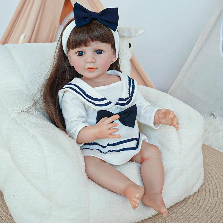 Reborn toddler doll in a sailor-style dress, sitting comfortably on a plush chair, holding a teddy bear and gazing gently forward.