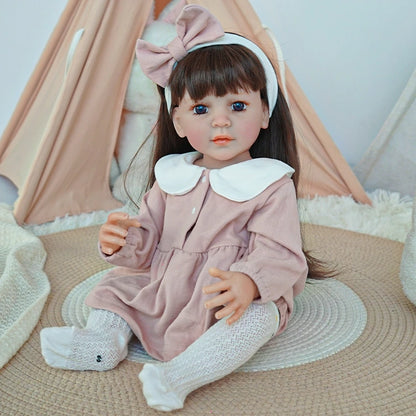 Reborn toddler doll sitting cross-legged on a rug, wearing a pink outfit with a large bow and white socks.