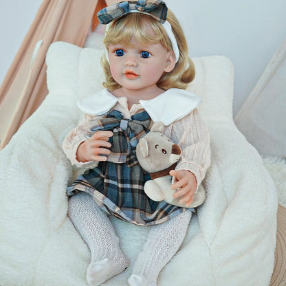 Reborn toddler doll sitting on a plush chair, wearing a plaid dress with a white collar, holding a small teddy bear.