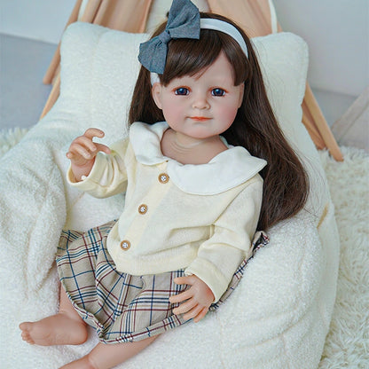 Reborn toddler doll sitting on a plush chair, wearing a cream top with a plaid skirt and a blue bow in her hair.