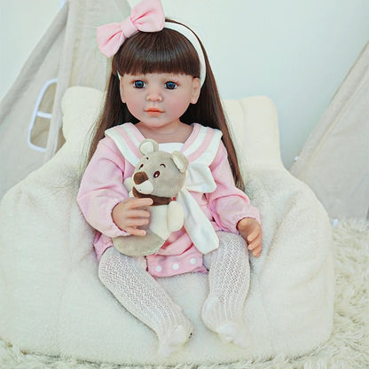 Reborn toddler doll in a pink dress and white bow, sitting on a plush chair with a small stuffed bear.