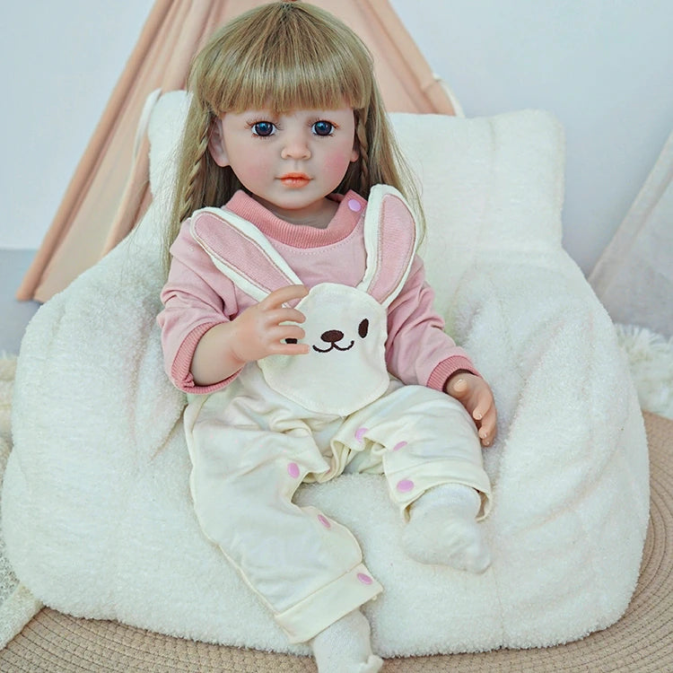 Reborn toddler doll sitting on a plush chair, dressed in pink and white, holding a teddy bear in her arms.