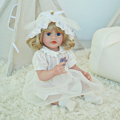 Reborn toddler doll sitting on a soft rug, dressed in a white dress with a matching bonnet, looking forward.