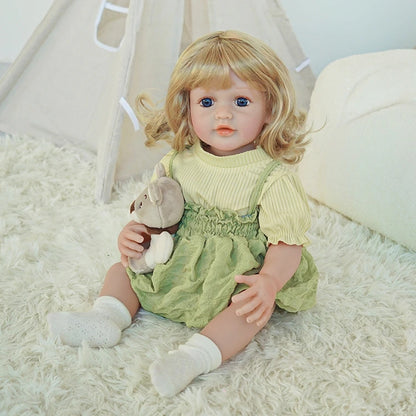 Close-up of reborn toddler doll wearing a green dress and holding a teddy bear, with her blonde hair styled in soft curls.