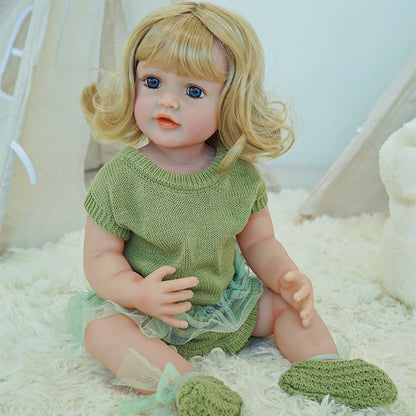 Reborn toddler doll in a green outfit, sitting on a soft white surface with a plush toy and shoes nearby.