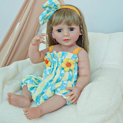 Reborn toddler doll sitting on a soft surface, dressed in a colorful romper with a blue bow in her hair, holding a plush toy.