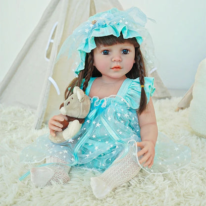 Reborn toddler doll sitting on a soft surface, dressed in a blue dress and bonnet, holding a teddy bear.