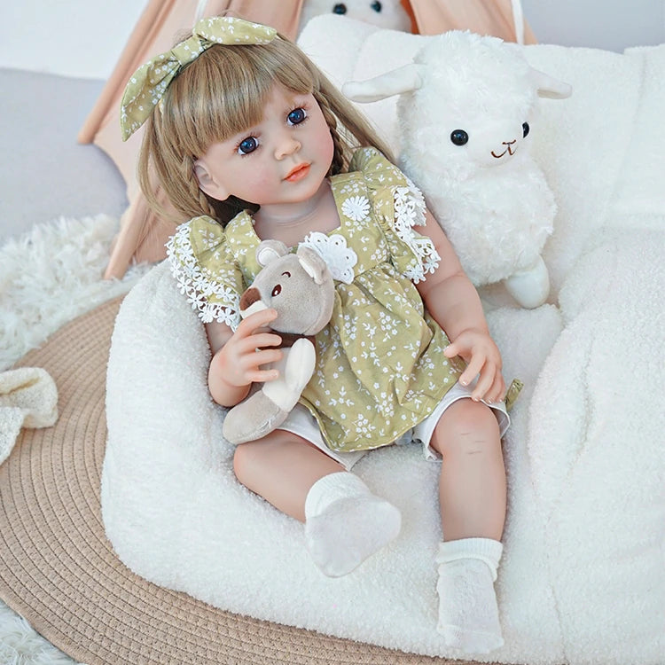 Reborn toddler doll sitting on a soft chair, wearing a green dress with lace accents, holding a teddy bear and looking to the side.