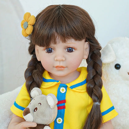 Close-up of reborn toddler doll with a yellow flower in her hair, wearing a bright yellow dress with blue accents and holding a teddy bear.