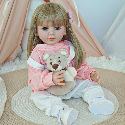 Reborn toddler doll sitting on a rug, dressed in a pink and white outfit, holding a teddy bear, with a flower clip in her hair.