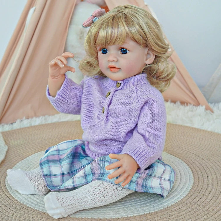 Reborn toddler doll sitting on a soft surface, wearing a purple sweater and plaid pants, looking forward with her hand raised.