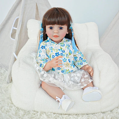  Reborn toddler doll sitting on a plush surface, dressed in a blue and white floral outfit, holding a small teddy bear.