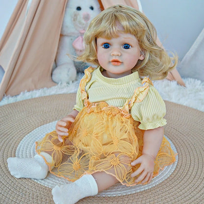 Reborn toddler doll sitting on a soft rug, wearing a yellow dress with floral patterns and white socks, holding a teddy bear.