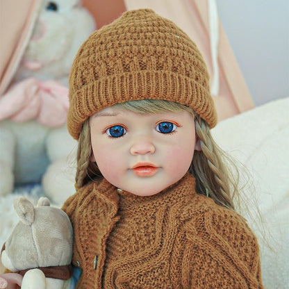 Close-up of a reborn toddler doll with blonde hair, wearing a brown knit outfit and holding a small teddy bear.