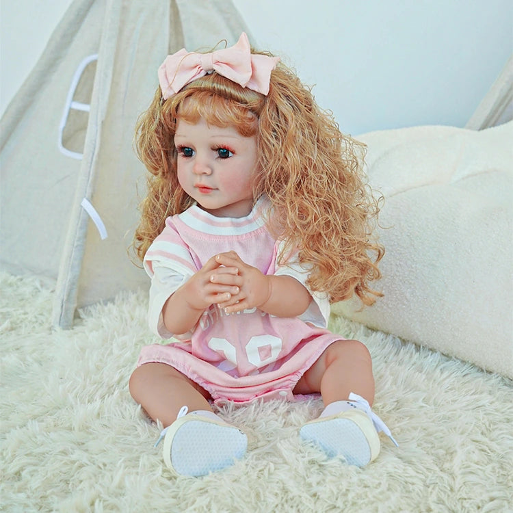 Reborn toddler doll with hands resting in her lap, wearing a cute pink dress and matching bow, sitting on a fluffy rug.