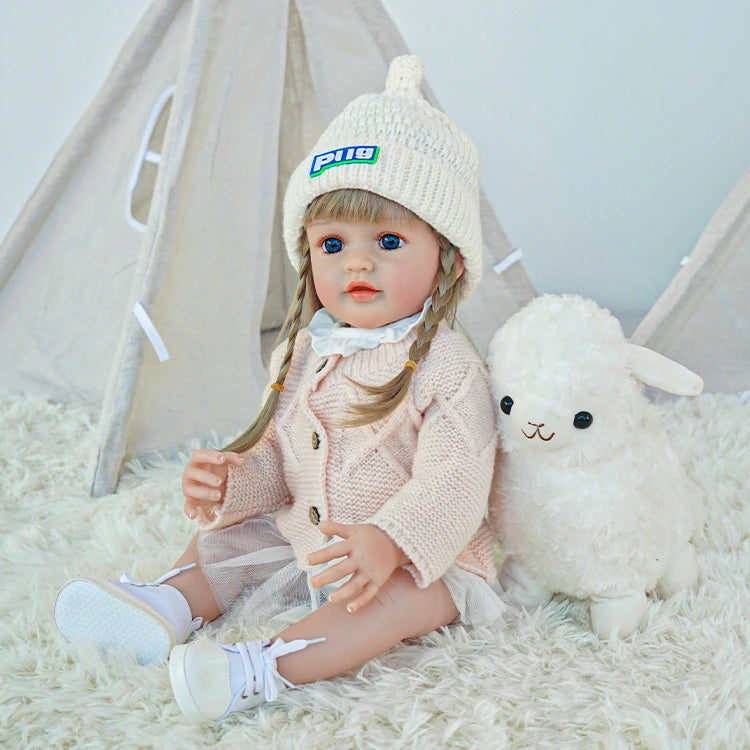 Reborn toddler doll sitting on a soft blanket, dressed in a pink coat and holding a stuffed animal.