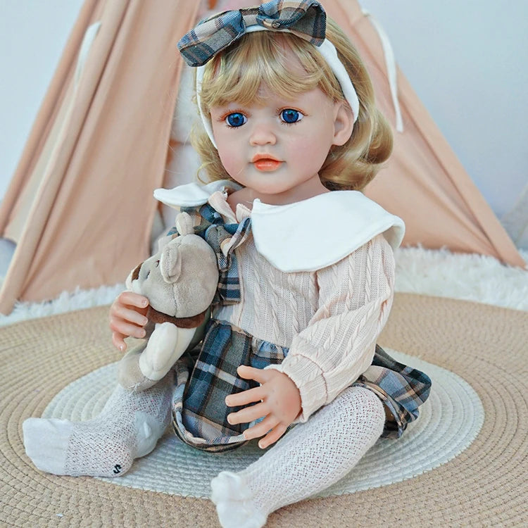 Reborn toddler doll sitting on a soft rug, dressed in a plaid dress with white tights, holding a teddy bear with a gentle expression.