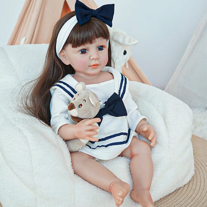 Close-up of reborn toddler doll sitting on a plush chair, dressed in a sailor outfit and holding a small plush teddy bear.