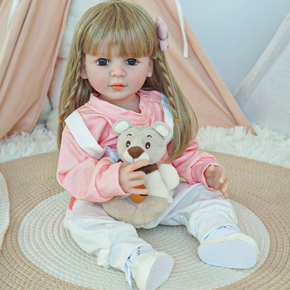 Reborn toddler doll sitting on a rug, wearing a pink sweater and white overalls, holding a teddy bear.