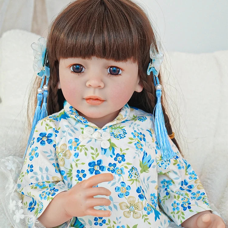 Close-up of reborn toddler doll with blue hair ribbons, wearing a floral blue top, looking forward with a gentle expression.