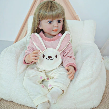 Reborn toddler doll sitting in a soft chair, dressed in white bunny-themed overalls and a pink top, holding a small plush toy.