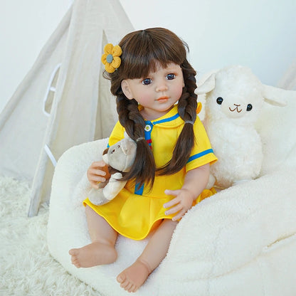 Reborn toddler doll sitting on a plush surface, dressed in a yellow dress with blue details, holding a teddy bear, looking to the side.