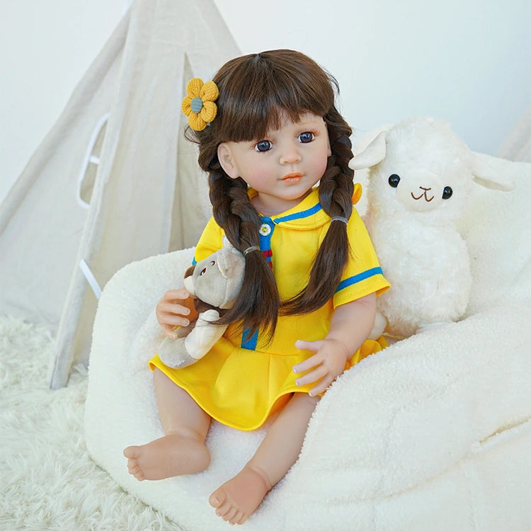 Reborn toddler doll sitting on a plush surface, dressed in a yellow dress with blue details, holding a teddy bear, looking to the side.