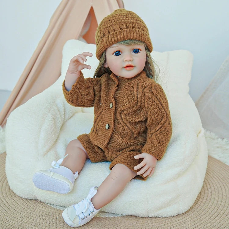 Reborn toddler doll sitting on a soft surface, lifting her hand while wearing a brown knit outfit and white shoes.