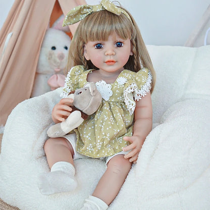 Close-up of reborn toddler doll sitting on a plush chair, wearing a green dress with lace sleeves, holding a small teddy bear.