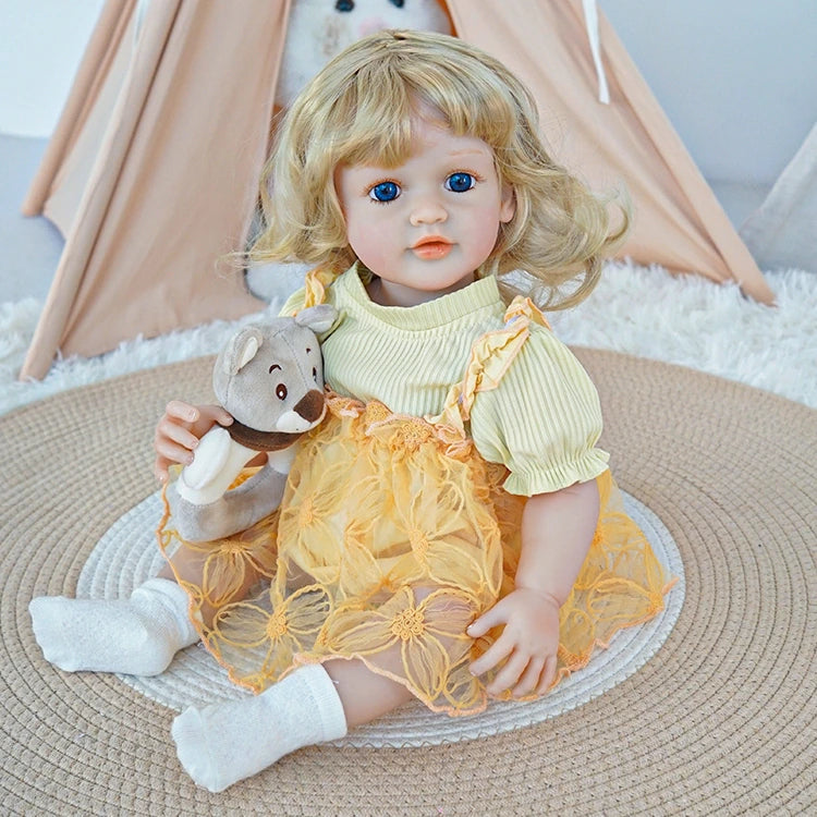 Reborn toddler doll sitting on a soft surface, wearing a yellow dress with floral details, holding a teddy bear, with a gentle expression.