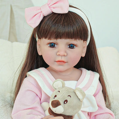 Close-up of reborn toddler doll with long straight brown hair, wearing a pink outfit and holding a plush toy.