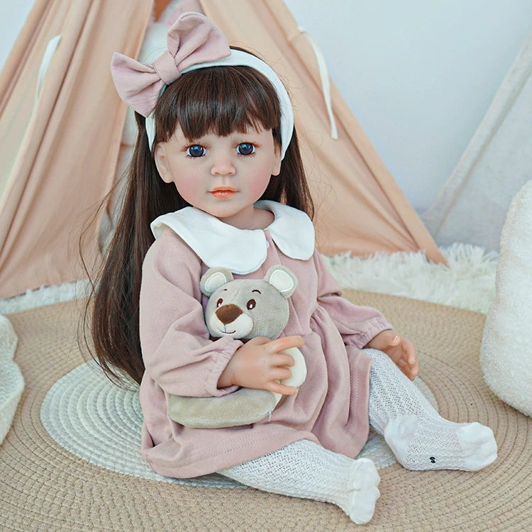  Reborn toddler doll dressed in a pink dress and white tights, sitting on a soft rug with her teddy bear.