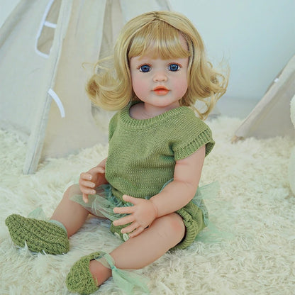 Reborn toddler doll sitting with crossed legs, wearing a green outfit and matching knit shoes.