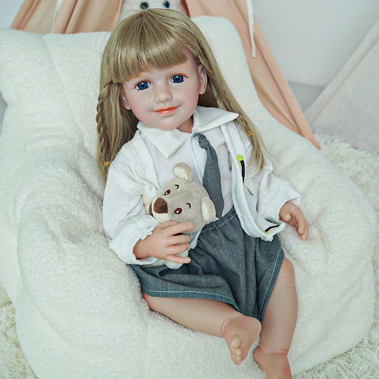 Reborn toddler doll sitting on a soft chair, holding a teddy bear, wearing a white shirt and gray skirt.