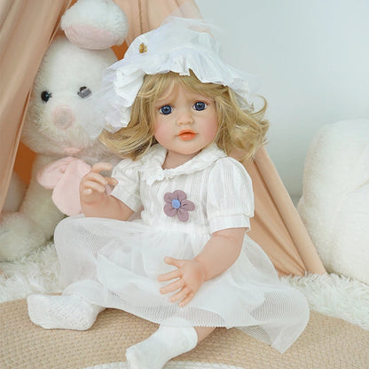 Reborn toddler doll sitting on a plush surface, dressed in a white dress with floral accents and a matching bonnet.