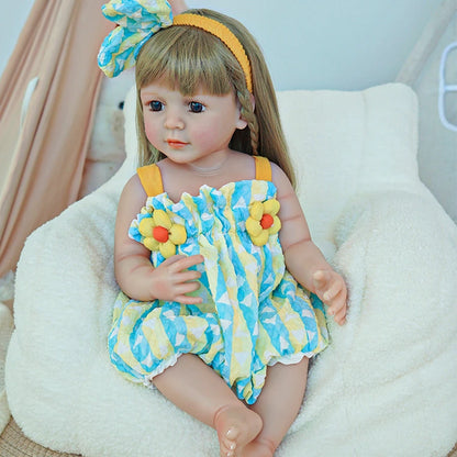 Reborn toddler doll sitting on a soft chair, wearing a blue and yellow outfit with floral details, holding a teddy bear.