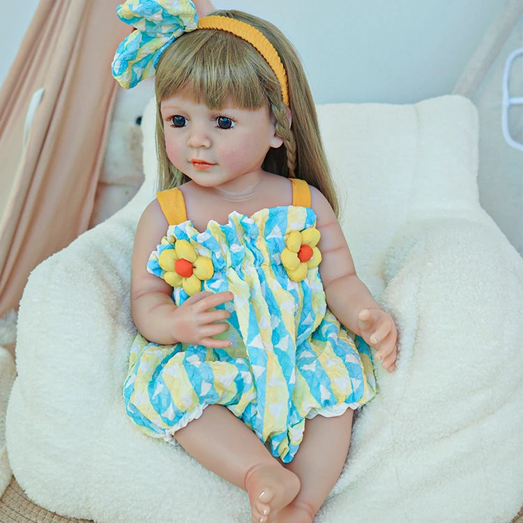 Reborn toddler doll sitting on a soft chair, wearing a blue and yellow outfit with floral details, holding a teddy bear.