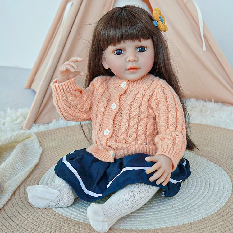 Reborn toddler doll raising one hand while sitting on a rug, dressed in a peach sweater and navy blue skirt.