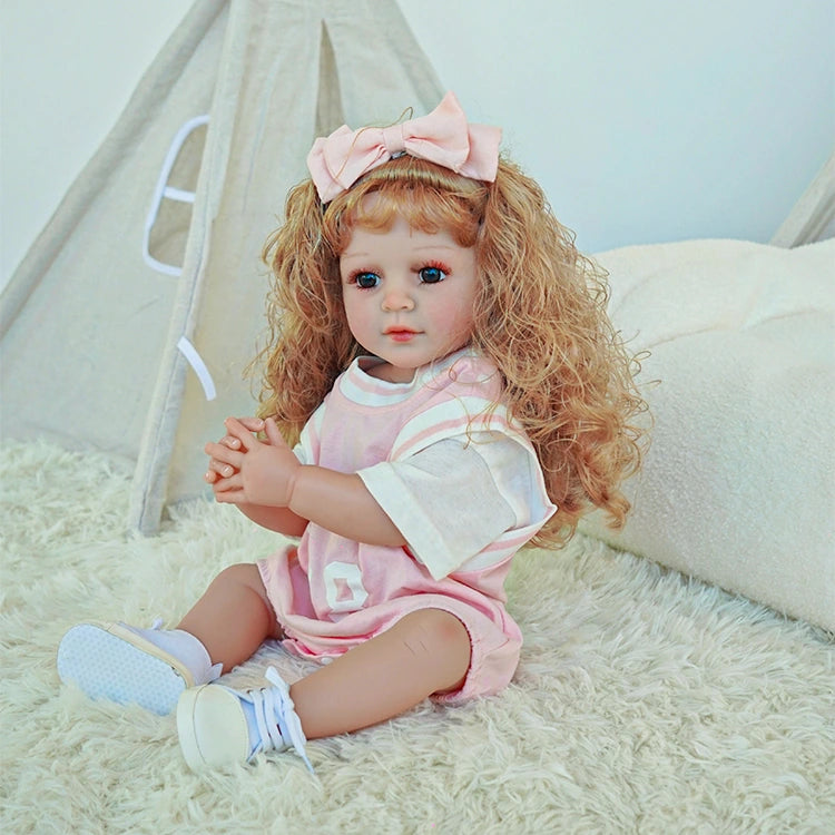 Reborn toddler doll sitting cross-legged, wearing a pink outfit and a large bow in her curly hair.