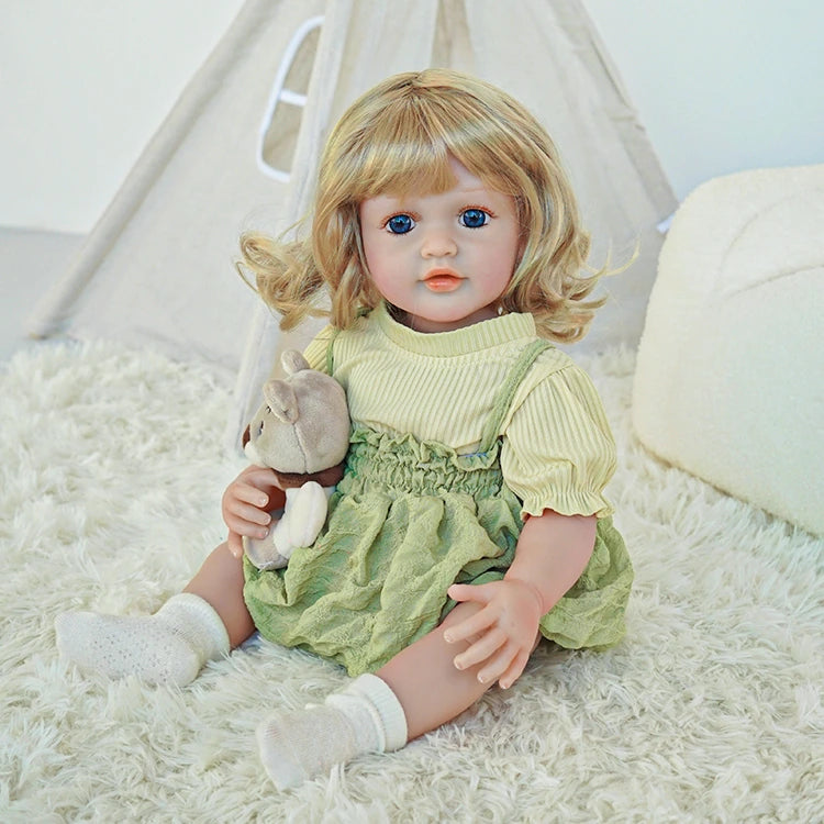 Reborn toddler doll sitting on a soft surface, wearing a green outfit and holding a teddy bear, looking slightly to the side.