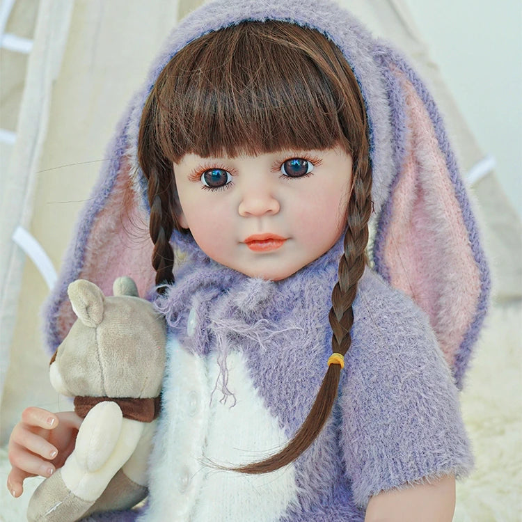 Reborn toddler doll with long bunny ears and braids, holding a small teddy bear while wearing a purple fluffy outfit.